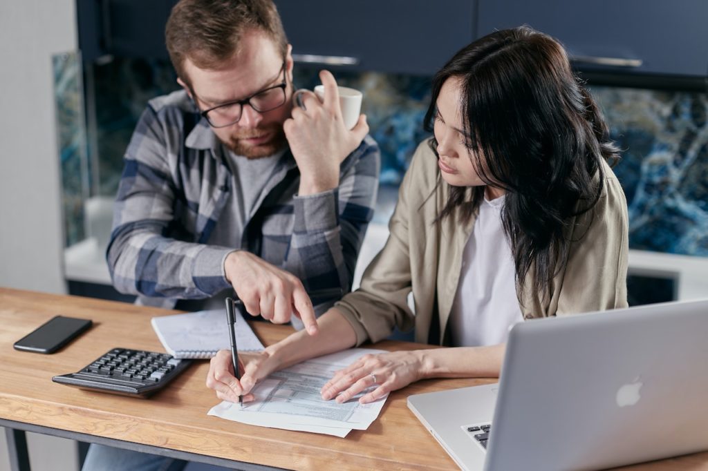 Couple calculating all their bills. The woman fills in some data on a paper. There is a calculator, a laptop and a mobile phone on the table as well.